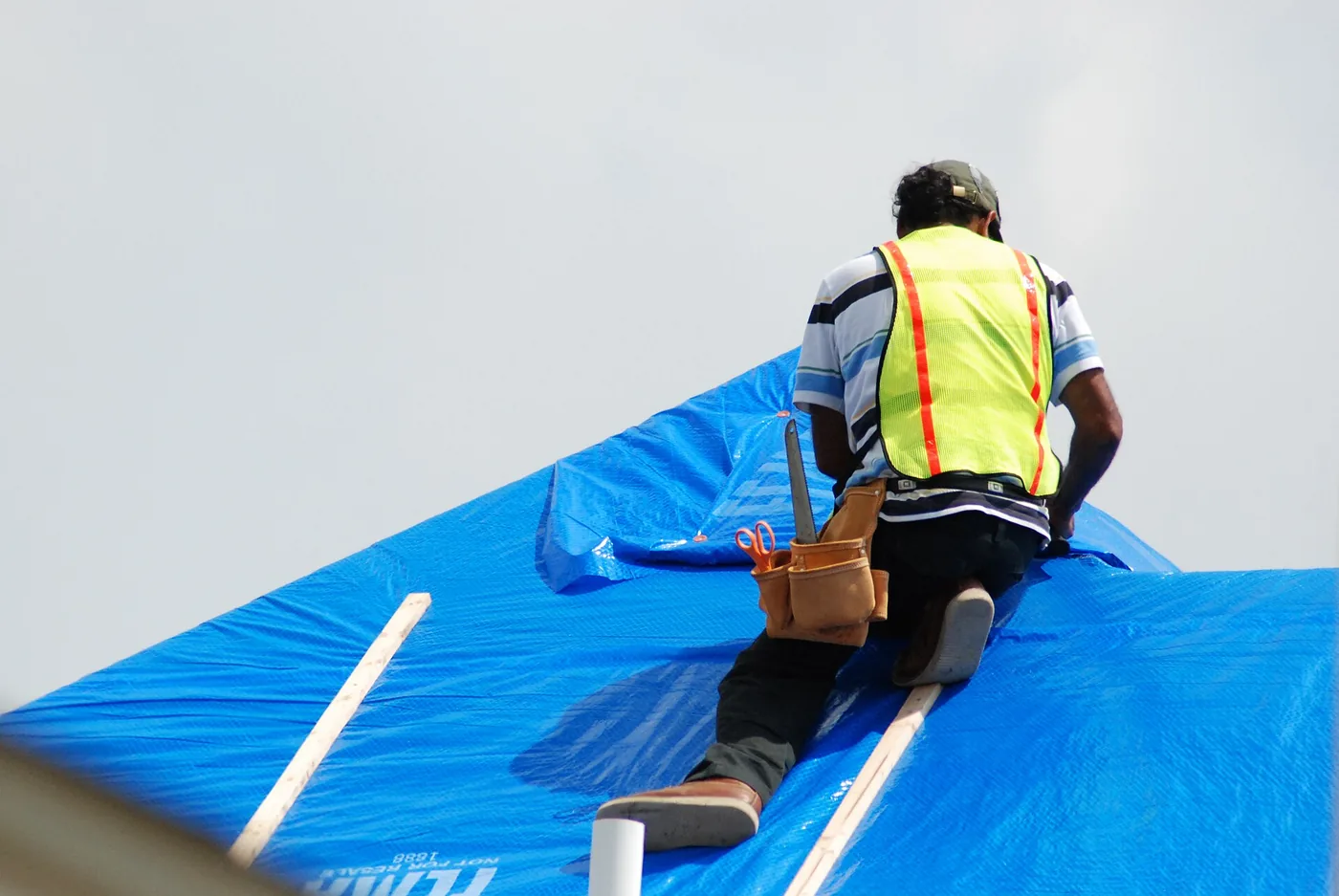 Roofing crew member securing a blue tarp with wood battens on a storm-damaged roof — Escambia County emergency tarping